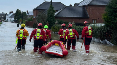 A group of men in red jackets and yellow helmets pulling a red boat through a flooded street with a number of houses in the background.