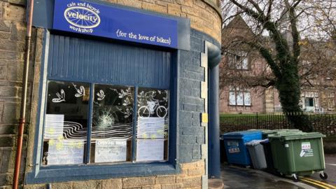 The exterior of the business has a blue frontage. A sign above a window includes the words: "For the love of bikes". There are industrial wheelie bins on a corner of the street next to the building.