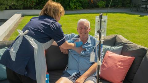 A nurse administers treatment while Des sits and smiles on his sofa in the garden