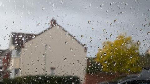 Rain drops on the outside of a window, captured from indoors. The droplets are in sharp focus. The side of a house and tree in the background are in soft focus