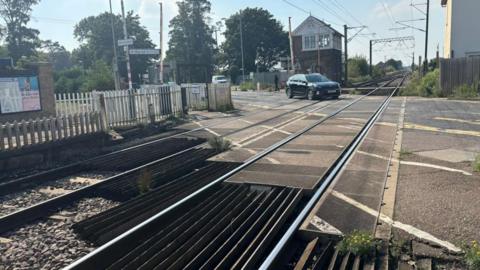 Level crossing at Foxton during the day. The gates are open and a black car is driving across the train tracks. There are markings on the road and to the side of it where there is a gate for pedestrians to cross. 