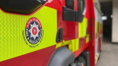 The side of a red-and-yellow fire engine bears the logo of the Isle of Man Fire and Rescue Service.