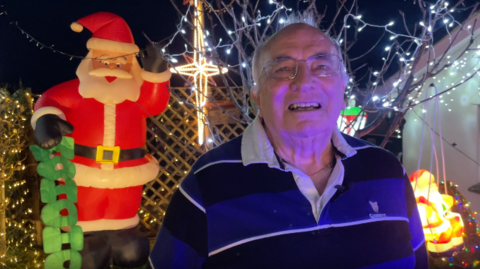 Mr Langman is stood in his garden which has been decorated with Christmas lights. Mr Langman is wearing a black and blue striped Guinness shirt. He has short hair and wears glasses. He is stood in front a small tree which has been decorated with fairy lights. A large inflatable Father Christmas decoration has been placed behind Mr Langman.