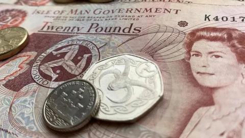 A close-up of an Isle of Man £20 note, with the head of Queen Elizabeth II and the three legs of Mann, and three coins placed on top of it. 