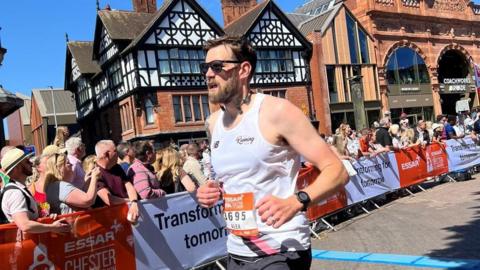 Alex Trivino with short dark hair and beard wearing white running vest with number 3695 running past historic houses with spectators behind barriers