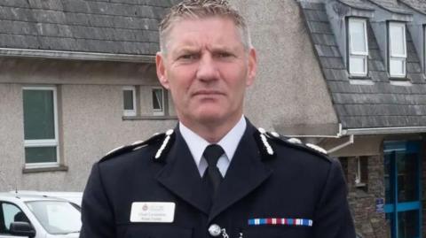 Russ Foster looking sternly at the camera. He is wearing his black uniform and name badge and standing in front of police headquarters in Douglas, with is a grey building with a glass sliding door with a blue frame with a car parked outside.