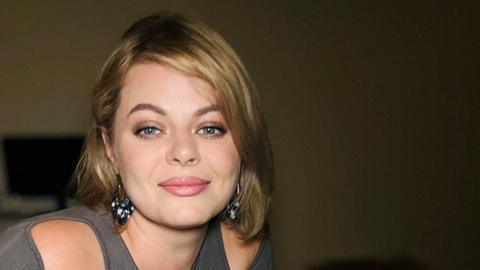 A young woman smiles into the camera. She has large green eyes and mid-length hair that falls onto her shoulders. She is wearing large dark earrings and a grey top which reveals her shoulders. 