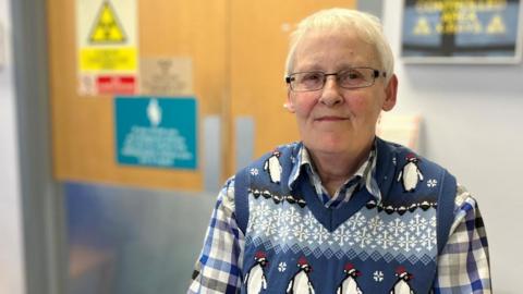 Dianne Covey pictured at a Royal Surrey NHS facility wearing a blue and white checked undershirt and a woollen pullover featuring penguins and snowflakes. The hospital setting in the background has been blurred.