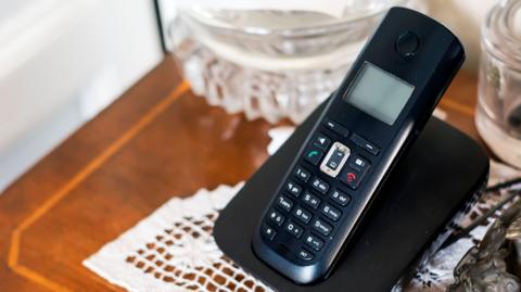 A black landline phone, switched off, in its charging stand. The charging stand is on a brown table, which had a white doily on it. There are two glass bowls, one to the left of the phone and on to the right. 