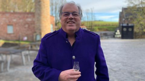 A man with glasses and grey hair in a blue top holds a small whisky glass in his hands outside a distillery building