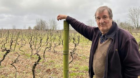 Winemaker George in his vineyard in Leeds, leaning against a wooden post and wearing a blue fleece, brown knitted jumper and blue and red checked shirt. He has brown hair and is 78 years old.