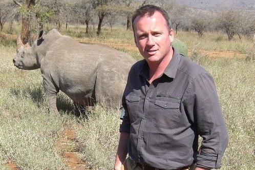 Martin Geisler in grey shirt with Rhino in background and thin trees and dry grassland around.