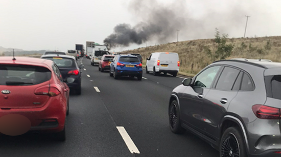 The M6 lined with cars across three lanes. Black smoke can be seen rising in the distance.
