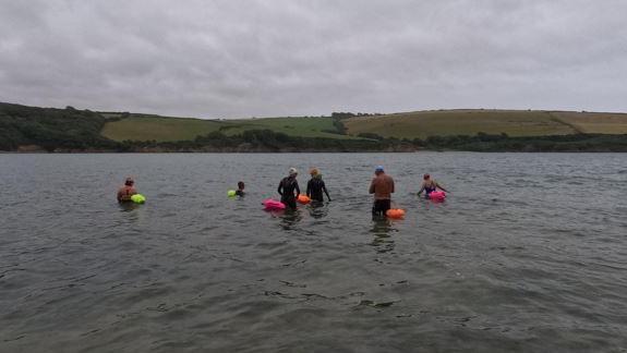 Swimmers in a river with colourful floatation devices and a river bank in the distance.