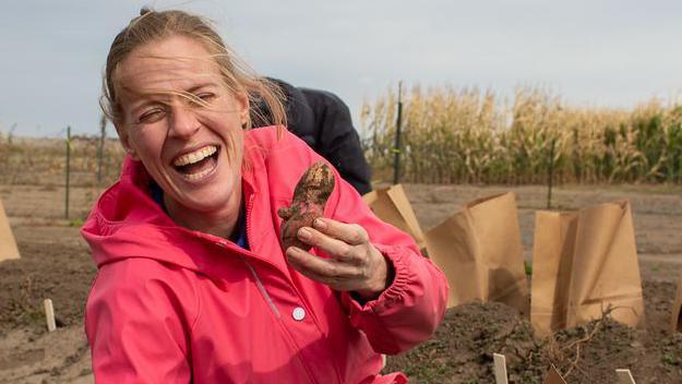 Dr Katherine Meacham-Hensold is laughing while holding a potato in a muddy field. She has blonde hair which has been blown across her face and she is wearing a pink jacket.