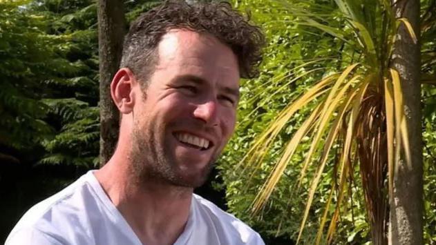 Mark Cavendish smiling. he's wearing a white T-shirt and sitting in a garden with green trees behind him.