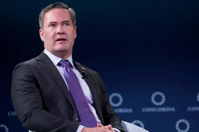 Mike Walts sits in a seat during a talk. He wears a purple tie and the background is blue.