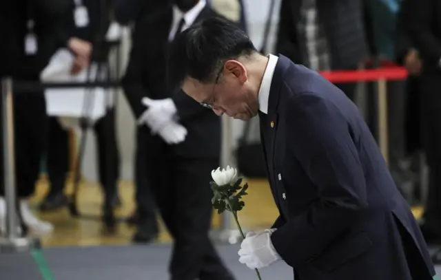 The acting president in a black suit and white gloves. He holds a white flower and bows