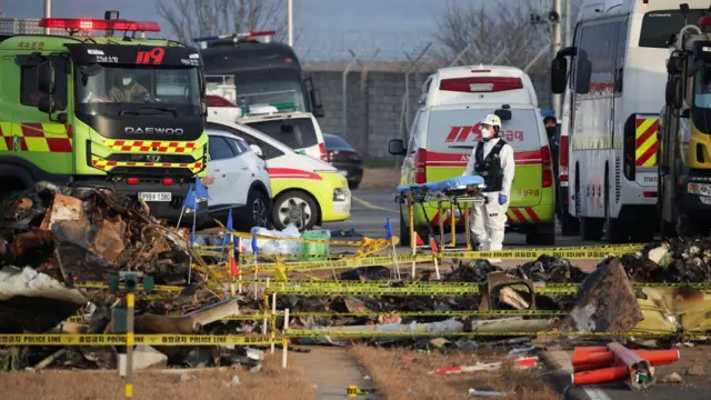 A man in a white suit stands next to a stretcher as debris from the plane lies in front of him, covered in police tape, a firefighter sits in a fire engine on the left