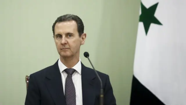 Bashar al-Assad sitting down in black suit, white shirt and patterned tie with a table mic in front of him. The Syrian flag (red top, white centre with two green stars and black bottom) is to his left