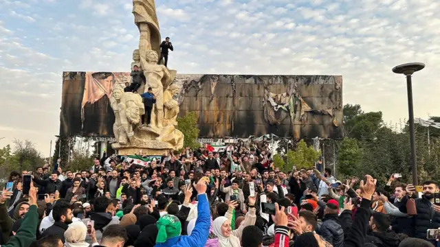 Hundreds of people pack a square in Syria celebrating the end of the Assad regime. Many hold their phones in the air filming the scene