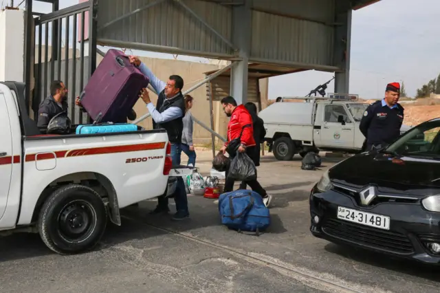 Man putting luggage into the back of a ute and others carrying bags. A uniformed officer is on the right