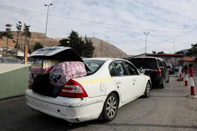 Line of cars at a border crossing. Car closest to camera has the boot overflowing with large plastic carrier bags with the boot unable to close. Mountains in the background