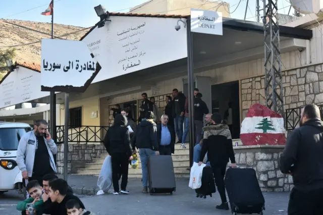 People carrying suitcases gather outside an office at the Masnaa border crossing, with signs in Arabic and one sign with the word Depart and a rock painted with the Lebanese flag