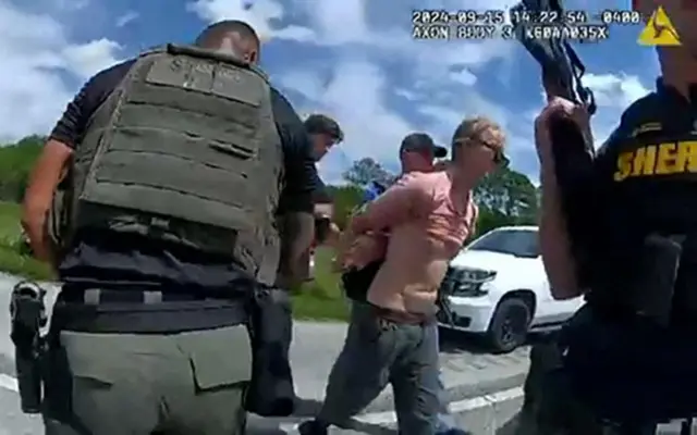 Screengrab from bodycam footage when Ryan Routh was arrested. Shows a man in a pink t-shirt with his arms held behind him by police on a road during the day. A man holding a gun is in the foreground.