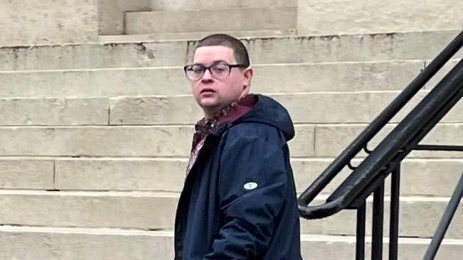 Ieuan Crump wearing a long navy coat and dark glasses, standing in front of steps