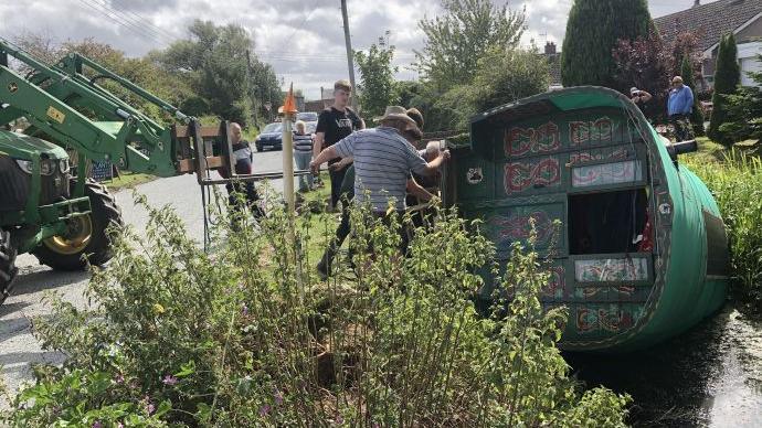 An old traditional style wagon turned on its side in a ditch being lifted out by people