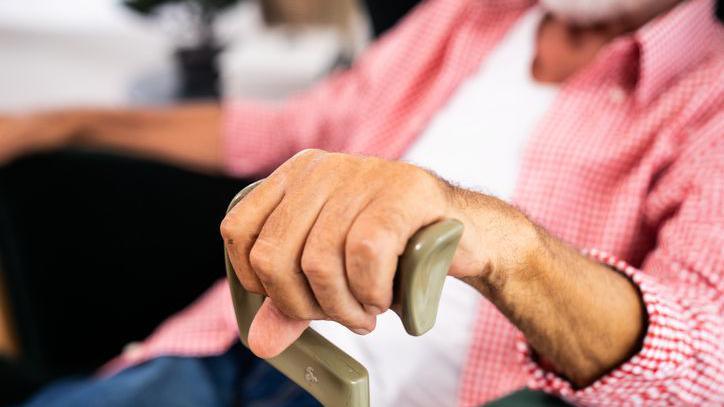 An older person, wearing jeans and a red and white checked shirt with a white top, is holding a walking cane and sitting in a chair.