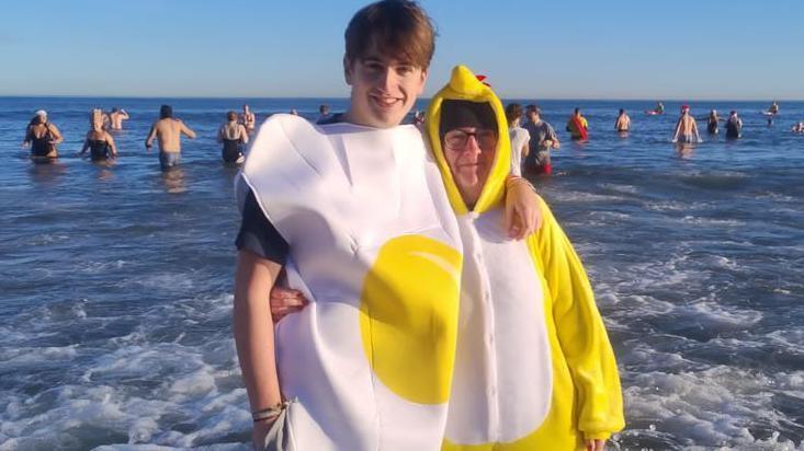 Dippers in yellow and white costumes embrace in the water. There are dozens behind them up to their waists in water. 