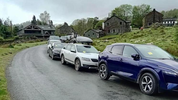 A line of cars by the side of an open rural road with yellow parking tickets in front of a number of stone cottages.