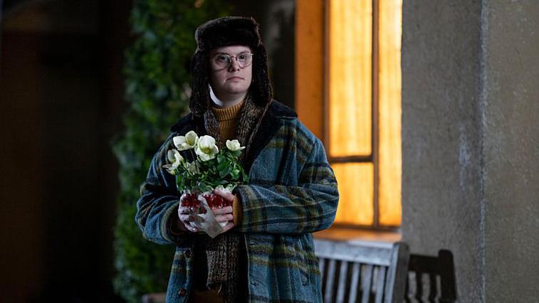 Reggie Jackson, played by Daniel Laurie, holding flowers outside in a hat and coat