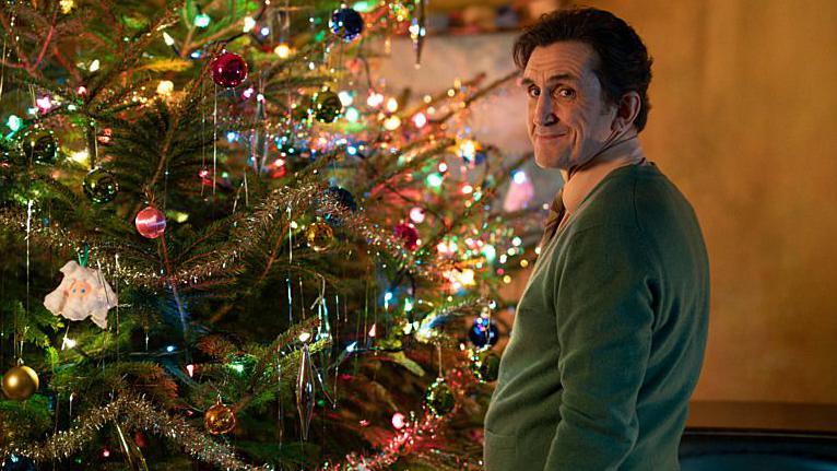 Stephen McGann as Dr Turner, standing by a brightly decorated Christmas tree in a green jumper