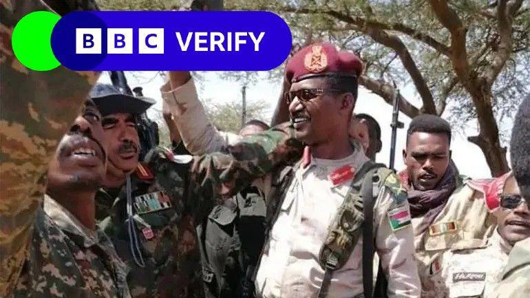 Former RSF commander Abu Aqla Keikal seen surrounded by troops. He holds a firearm aloft and is wearing a uniform and sunglasses. The men surrounding him are also wearing uniforms. 