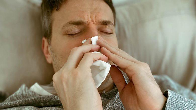 Man with stubble wearing grey top, blowing nose with a white tissue.