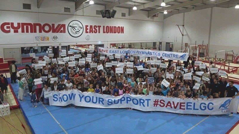 A large group of people, including children and adults, gather in a gym holding signs in support of saving the East London Gymnastics Centre. They stand behind a banner reading, "SHOW YOUR SUPPORT #SAVEELGC." The gym has "East London" and "Gymnova" signage in the background.