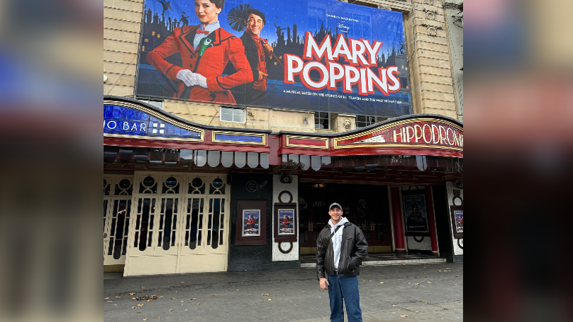 Rhys Batten standing outside the front of the Bristol Hippodrome Theatre, with a poster for the show on the wall behind him