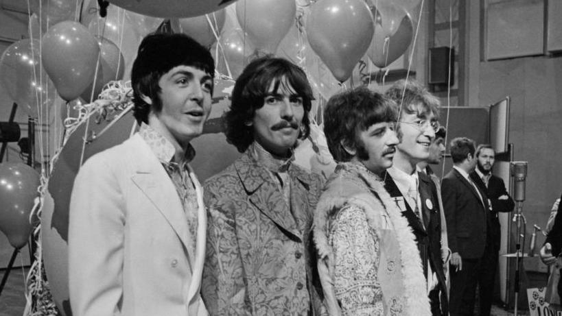 The Beatles are pictured lined up together during press call at Abbey Road Studios in a black and white photo. There are balloons behind them and music recording equipment. They are wearing jackets and multi-patterned shirts.