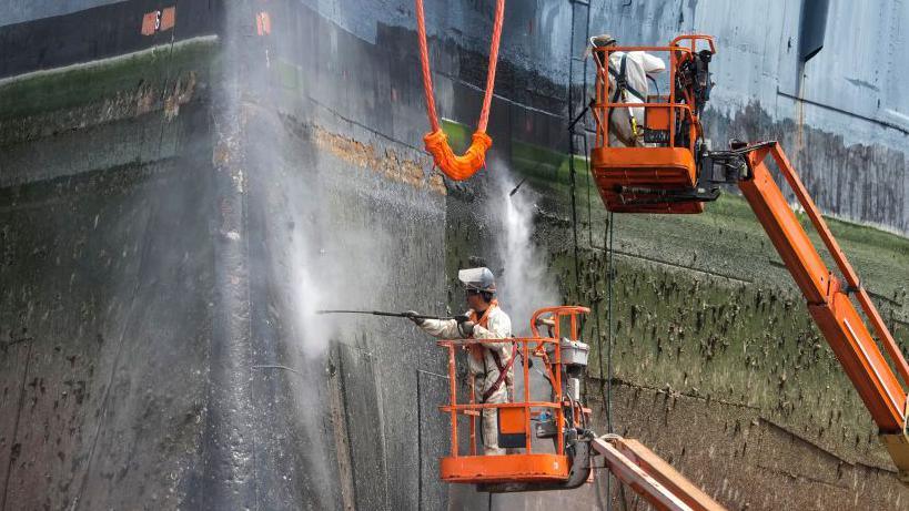 Workers clean the hull of a large ship with pressure hoses