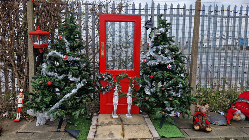 A red door with a wreath is sandwiched between two christmas trees decked with tinsel.