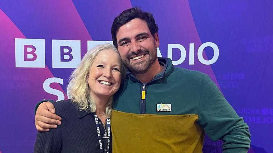 A man with short dark hair and dark stubble stands with his arm around a BBC presenter with long blonde hair. They are both smiling at the camera and standing in front of a purple backdrop with the words BBC Radio Solent on it