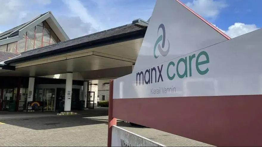 A modern entrance to a hospital, with a overhanging shelter above it and a glass steepled roof. In the foreground focus is a white triangular sign which reads "Manx Care" in grey and green font, below the healthcare service's celtic swirl logo. 