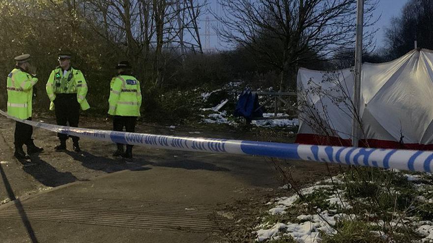 Three police officers, wearing yellow hi-vis jackets, stand behind police tape which cordons off the area in which a baby's remains were found. 