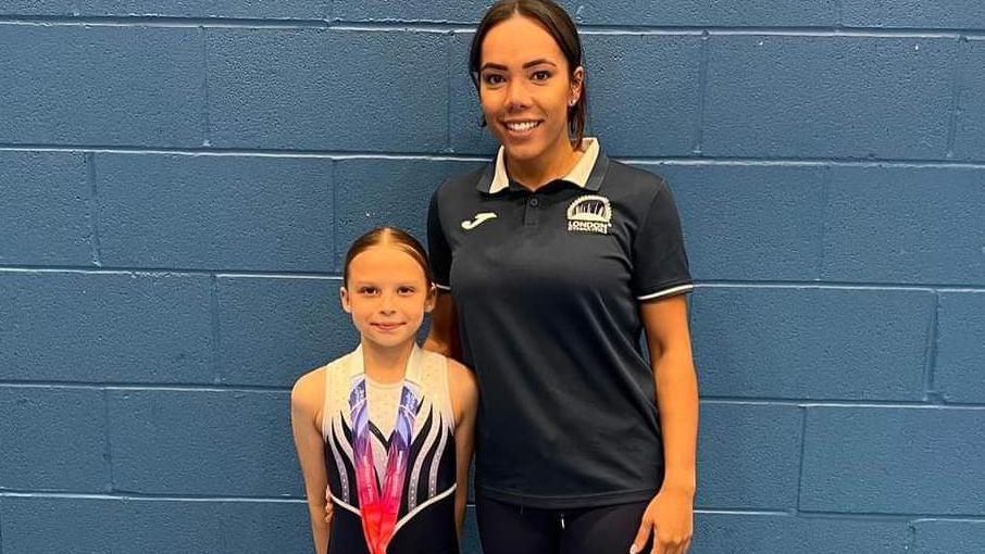 Mikayla Metcan standing with her coach. She is wearing a gymnastics leotard and has a medal around her neck. Her coach is wearing a black polo shirt.