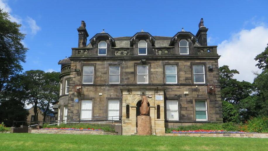 A three-storey residence built in the 1700s and featuring Georgian bow windows looms over a sloped lawn with blue skies above and trees seen on either side. Flower beds can be seen running across the front of the property on either side of a small statue. 