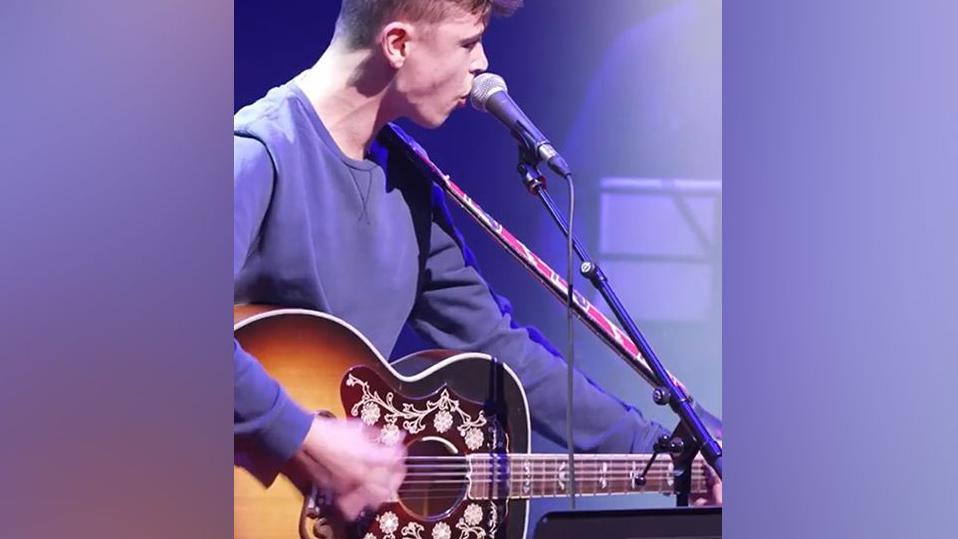 Side view of Tom A Smith playing a guitar, seen from waist height. He has short hair and is wearing a purple sweatshirt. 
