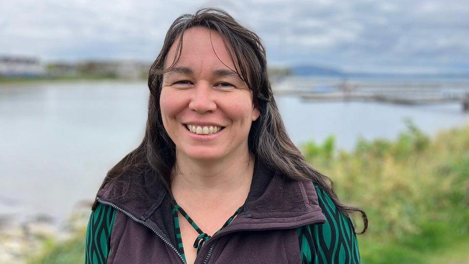 Marianne Green smiling at the camera on Rathlin Island.  She has long, dark hair and is wearing a dark-coloured bodywarmer over a green and black patterned top.